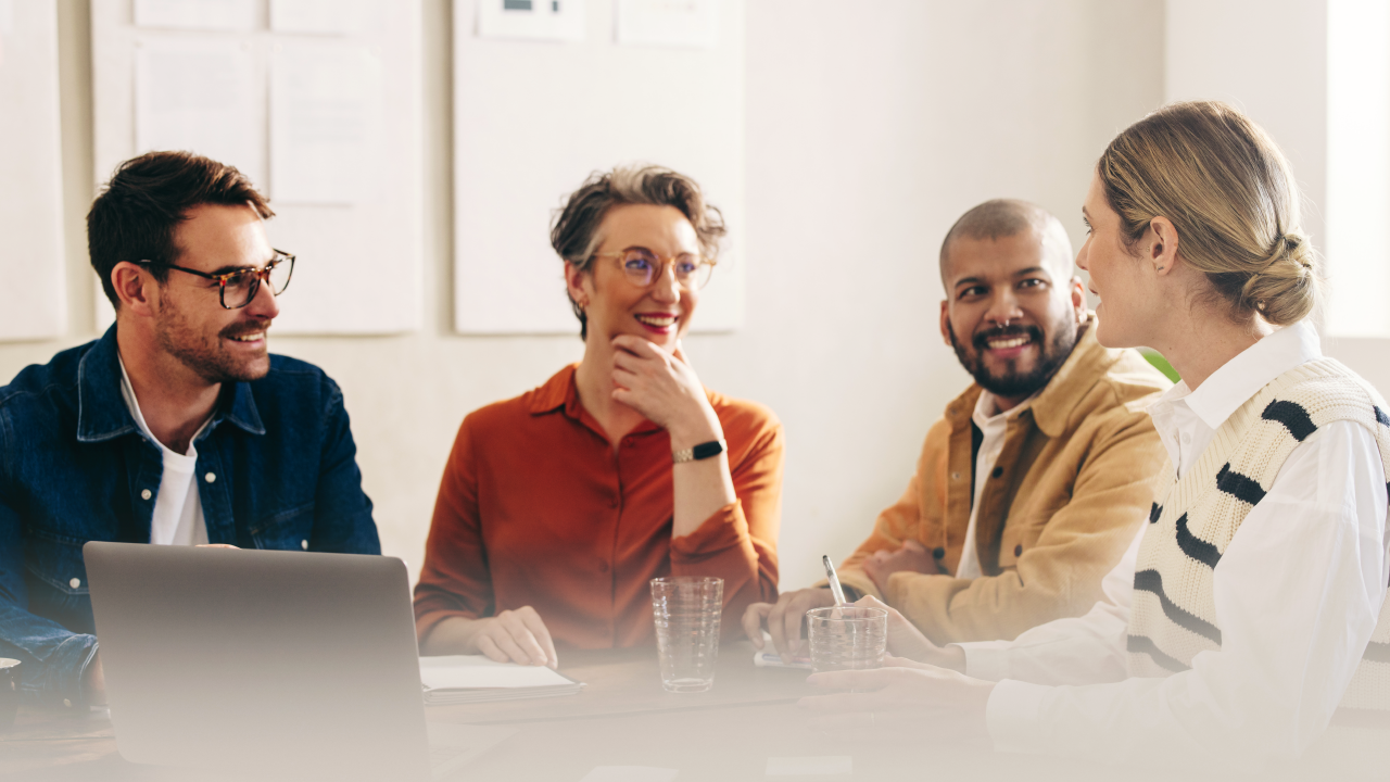 A group of coworkers laugh and converse seated at a table with a laptop